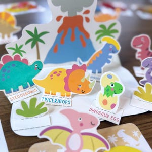 K&ouml;nnte beinhalten: Eine Sammlung bunter Dinosaurier-Papierausschnitte, darunter ein blaugr&uuml;ner Stegosaurus, ein gelber Triceratops und ein gr&uuml;ner Dinosaurier, der aus einem Ei schl&uuml;pft. Das Set enth&auml;lt einen zentralen Vulkanhintergrund, Palmen und Beschriftungen.