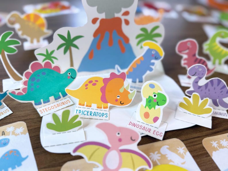 K&ouml;nnte beinhalten: Eine Sammlung bunter Dinosaurier-Papierausschnitte, darunter ein blaugr&uuml;ner Stegosaurus, ein gelber Triceratops und ein gr&uuml;ner Dinosaurier, der aus einem Ei schl&uuml;pft. Das Set enth&auml;lt einen zentralen Vulkanhintergrund, Palmen und Beschriftungen.