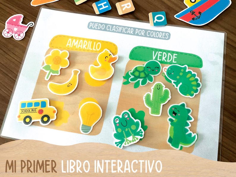 May include: Educational interactive book with colorful illustrations. Features yellow and green themed images, including a duck, banana, school bus, and a frog. Text reads "PUEDO CLASIFICAR POR COLORES" and "MI PRIMER LIBRO INTERACTIVO".