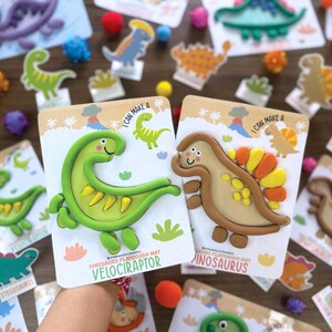 K&ouml;nnte beinhalten: Zwei Knetmatten mit Dinosaurier-Motiven, die einen gr&uuml;nen Velociraptor und einen beigen Spinosaurus zeigen. Jede Karte enth&auml;lt den Text I CAN MAKE A und den Namen des Dinosauriers sowie Illustrationen von Vulkanen und Palmen.