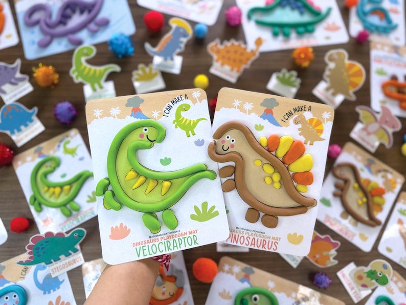 K&ouml;nnte beinhalten: Zwei Knetmatten mit Dinosaurier-Motiven, die einen gr&uuml;nen Velociraptor und einen beigen Spinosaurus zeigen. Jede Karte enth&auml;lt den Text I CAN MAKE A und den Namen des Dinosauriers sowie Illustrationen von Vulkanen und Palmen.