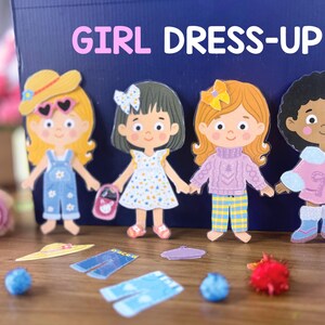 Pode incluir: Um conjunto de quatro bonecas de papel com v&aacute;rios trajes e acess&oacute;rios, exibido em frente a uma caixa azul-escura com a etiqueta GIRL DRESS-UP. As bonecas apresentam diversas cores e estilos de cabelo, vestindo roupas como jardineiras e vestidos.