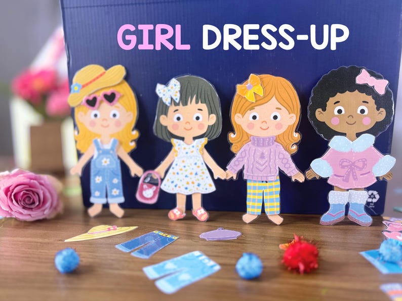 Pode incluir: Um conjunto de quatro bonecas de papel com v&aacute;rios trajes e acess&oacute;rios, exibido em frente a uma caixa azul-escura com a etiqueta GIRL DRESS-UP. As bonecas apresentam diversas cores e estilos de cabelo, vestindo roupas como jardineiras e vestidos.