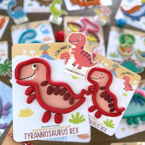 K&ouml;nnte beinhalten: Zwei Tyrannosaurus Rex Knetmatten mit einer rosa Dinosaurier-Illustration und roten Knet-Umrandungen. Die Karten enthalten den Text I CAN MAKE A TYRANNOSAURUS REX und zeigen eine Cartoon-Dinosaurier-Grafik mit Vulkan und Palmen.
