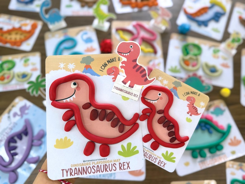 K&ouml;nnte beinhalten: Zwei Tyrannosaurus Rex Knetmatten mit einer rosa Dinosaurier-Illustration und roten Knet-Umrandungen. Die Karten enthalten den Text I CAN MAKE A TYRANNOSAURUS REX und zeigen eine Cartoon-Dinosaurier-Grafik mit Vulkan und Palmen.