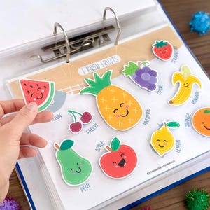Op de afbeelding: Een ordnerpagina met de titel I KNOW FRUITS met kleurrijke cartoon fruitstickers, waaronder een ananas, banaan, sinaasappel, citroen, appel, peer, kers, druif, aardbei en een vastgehouden watermeloenpartje, elk met een lachend gezichtje en label.