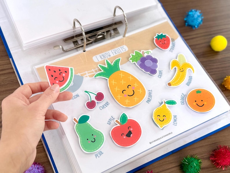 Op de afbeelding: Een ordnerpagina met de titel I KNOW FRUITS met kleurrijke cartoon fruitstickers, waaronder een ananas, banaan, sinaasappel, citroen, appel, peer, kers, druif, aardbei en een vastgehouden watermeloenpartje, elk met een lachend gezichtje en label.