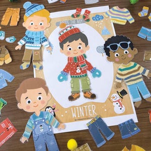 Pode incluir: Um conjunto de bonecos de papel intitulado DRESS THE BOY WINTER com tr&ecirc;s personagens de meninos ilustrados e v&aacute;rias pe&ccedil;as de vestu&aacute;rio de inverno, incluindo camisolas, gorros, cachec&oacute;is, luvas, cal&ccedil;as e sapatos, dispostos numa cena de inverno.