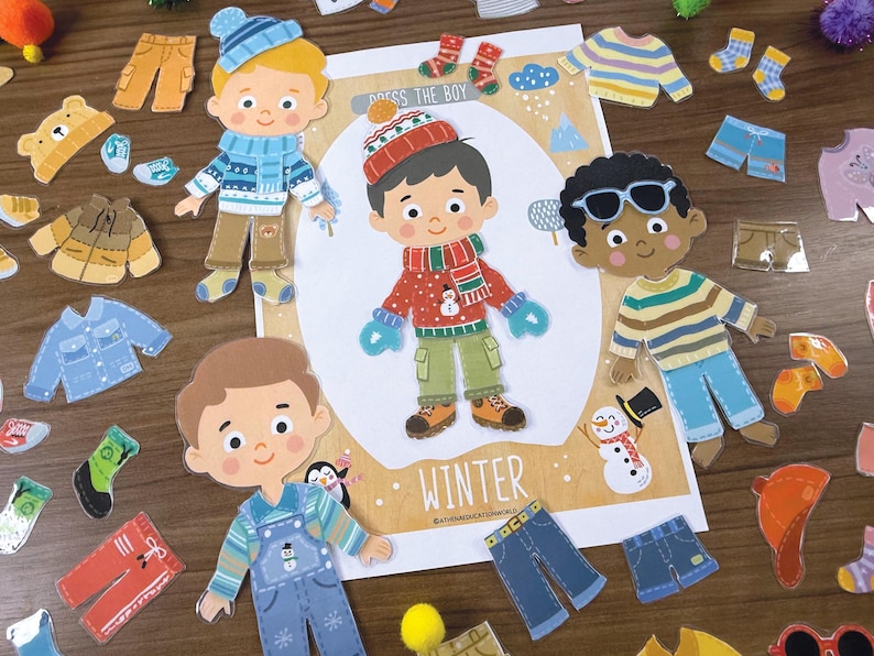 Pode incluir: Um conjunto de bonecos de papel intitulado DRESS THE BOY WINTER com tr&ecirc;s personagens de meninos ilustrados e v&aacute;rias pe&ccedil;as de vestu&aacute;rio de inverno, incluindo camisolas, gorros, cachec&oacute;is, luvas, cal&ccedil;as e sapatos, dispostos numa cena de inverno.