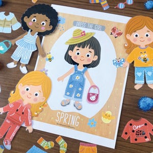 Pode incluir: Um conjunto de bonecas de papel intitulado Dress The Girl Spring com uma personagem central vestindo jardineiras jeans azuis com flores brancas, um chap&eacute;u de palha e uma bolsa rosa, rodeada por bonecas adicionais e v&aacute;rias pe&ccedil;as de roupa.