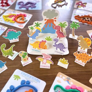 K&ouml;nnte beinhalten: Eine Sammlung von Bastelsets zum Thema Dinosaurier mit bunten Knetfiguren, Pappaufstellern verschiedener Arten wie Triceratops und Stegosaurus sowie einem zentralen Vulkan-Display. Jedes Set enth&auml;lt illustrierte Karten mit Dinosauriernamen.