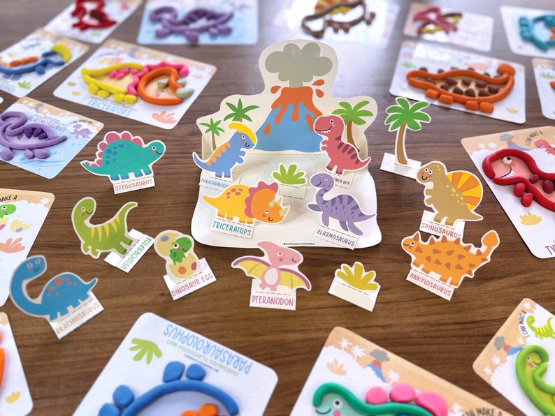 K&ouml;nnte beinhalten: Eine Sammlung von Bastelsets zum Thema Dinosaurier mit bunten Knetfiguren, Pappaufstellern verschiedener Arten wie Triceratops und Stegosaurus sowie einem zentralen Vulkan-Display. Jedes Set enth&auml;lt illustrierte Karten mit Dinosauriernamen.