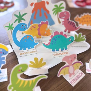 K&ouml;nnte beinhalten: Eine Sammlung bunter Dinosaurier-Papieraufsteller mit einem Vulkan-Hintergrund, Palmen und einzelnen Figuren mit den Beschriftungen Brachiosaurus, Stegosaurus, Parasaurolophus, Pteranodon und Dinosaur Egg in verschiedenen Blau-, Gr&uuml;n- und Rosat&ouml;nen.