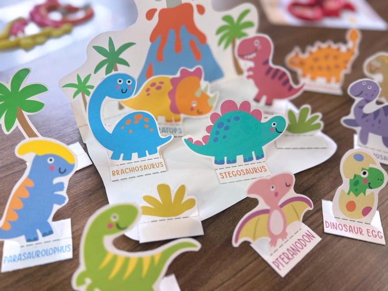 K&ouml;nnte beinhalten: Eine Sammlung bunter Dinosaurier-Papieraufsteller mit einem Vulkan-Hintergrund, Palmen und einzelnen Figuren mit den Beschriftungen Brachiosaurus, Stegosaurus, Parasaurolophus, Pteranodon und Dinosaur Egg in verschiedenen Blau-, Gr&uuml;n- und Rosat&ouml;nen.