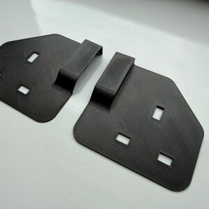 Puede incluir: Dos objetos de color gris oscuro, texturizados, de forma geométrica, con recortes rectangulares y un asa elevada. Los objetos están colocados sobre una superficie blanca y parecen estar hechos de un material compuesto.