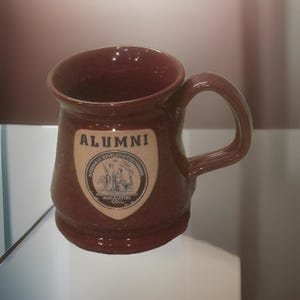 May include: A maroon ceramic mug with a curved handle. The mug features a cream-colored shield with the word "ALUMNI" at the top and an illustration below. The mug is a classic shape, suitable for hot beverages.