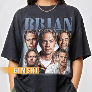 May include: Black t-shirt with a vintage-style graphic of Brian O'Conner from the Fast and Furious franchise. The graphic features multiple images of the character, including a close-up of his face. The text "Brian O'Conner" is printed in white above the graphic, and the text "GIMSKI" is printed in white below the graphic.
