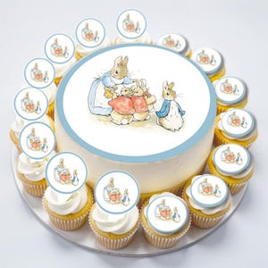 May include: A white cake decorated with a blue and white Peter Rabbit themed edible image. The cake is surrounded by 12 cupcakes, each topped with a round edible image of Peter Rabbit and his family.