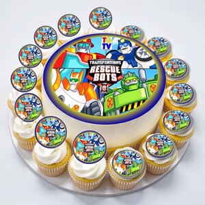 May include: A round cake and cupcakes decorated with a Transformers Rescue Bots theme. The cake has a blue border and features cartoon characters. Cupcakes are topped with edible images of the same theme. The cake and cupcakes are on a white plate.