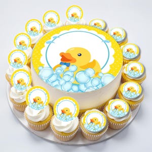 May include: A yellow rubber ducky cake topper with blue bubbles and a blue bow tie on a white cake with yellow polka dots. The cake is surrounded by cupcakes with matching toppers.