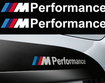 May include: A close-up of a black and white "M Performance" logo on a car's side skirt. The logo features the iconic BMW M tri-color roundel in blue, red, and white.