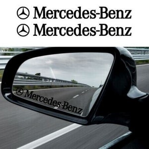以下が含まれることがあります： メルセデス・ベンツのロゴが入った「Mercedes-Benz」と書かれたステッカーが貼られた黒い車のサイドミラー。ミラーはガードレールのある道路を映し出している。