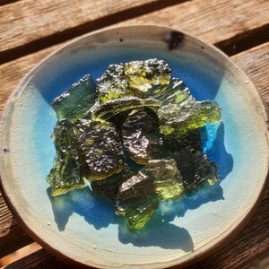 May include: A collection of rough, olive-green moldavite gemstones in a small, round, light blue and beige ceramic bowl. The gemstones have a textured, glassy appearance. The bowl is on a wooden surface.