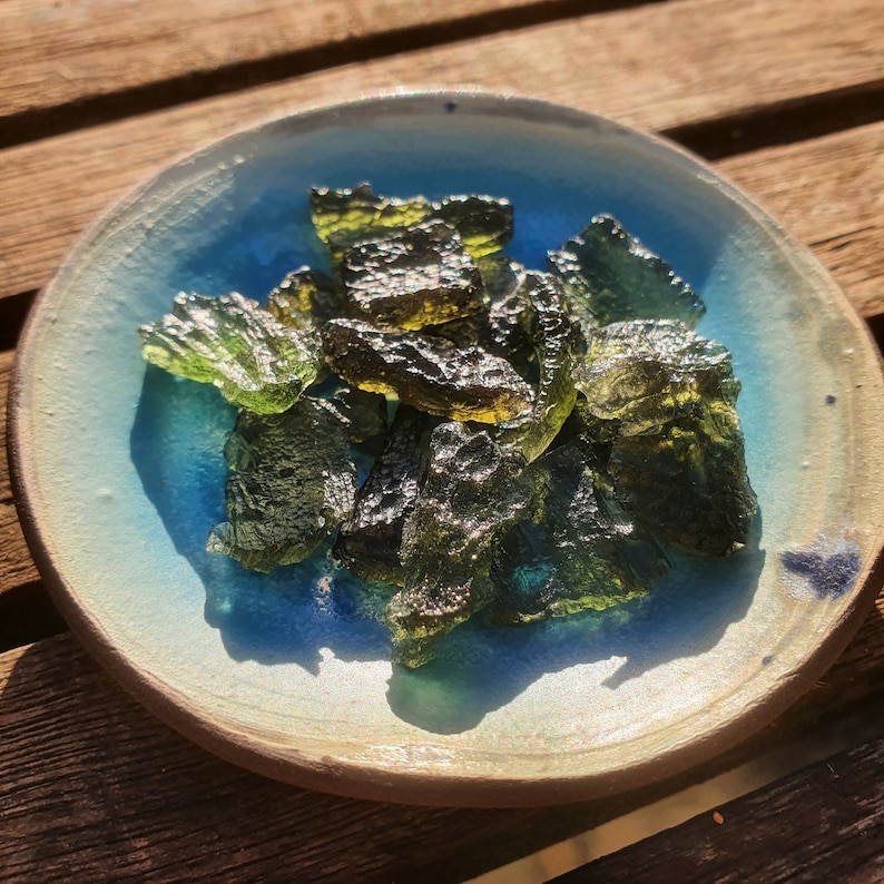 May include: A selection of raw, translucent green Moldavite stones presented in a small, round ceramic bowl. The bowl features a light blue interior and a brown rim. The stones exhibit a textured surface and varying shades of green.
