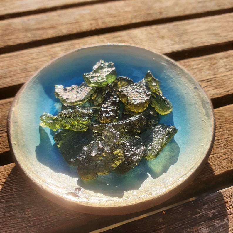 May include: A selection of translucent, olive-green moldavite crystals in a small, round ceramic bowl. The bowl features a light blue interior and a beige rim. The crystals have a rough, textured appearance.