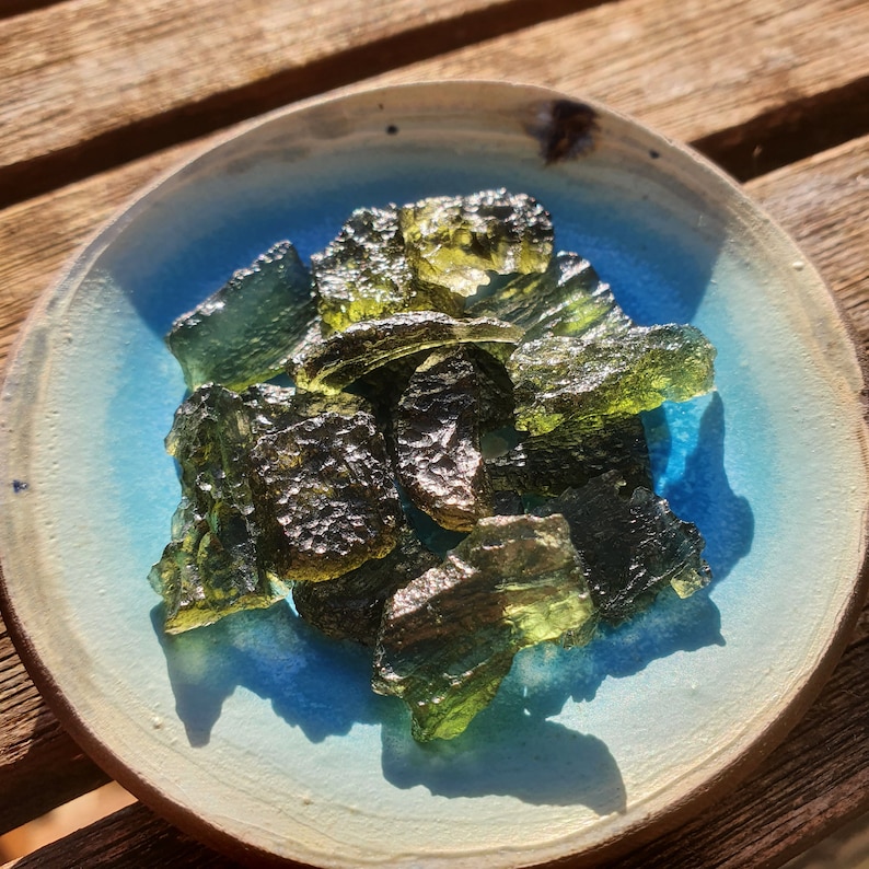 May include: A collection of rough, olive-green moldavite gemstones in a small, round, light blue and beige ceramic bowl. The gemstones have a textured, glassy appearance. The bowl is on a wooden surface.