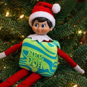 May include: A doll wearing a red and white Santa hat, a red long-sleeved outfit, and a white collar, holding a square, lime green and blue marbled Nee Doh fidget toy with the text Nee Doh embossed on the front surface.