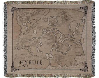 May include: A woven throw blanket featuring a detailed map of Hyrule in shades of beige and brown. The design includes geographical landmarks, mountain ranges, and the text Hyrule, all framed by a decorative border with fringed edges.