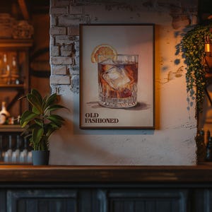 Op de afbeelding: Een ingelijste print van een Old Fashioned cocktail met ijsblokjes en een sinaasappelschijfje. De print hangt aan een bakstenen muur in een bar. De woorden "OLD FASHIONED" staan eronder gedrukt.