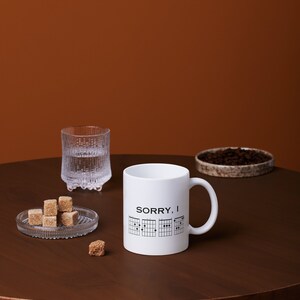 May include: White ceramic mug with a black graphic of guitar chords and the text "SORRY, I" on a brown wooden table with sugar cubes and a glass of water.