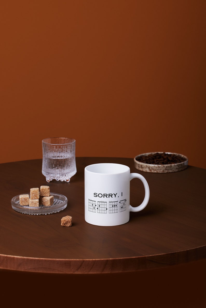 May include: White ceramic mug with a black graphic of guitar chords and the text "SORRY, I" on a brown wooden table with sugar cubes and a glass of water.