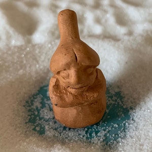 May include: A small, hand-crafted terracotta figurine of a person with a rounded head and a pointed hat. The figurine is standing on a white surface.