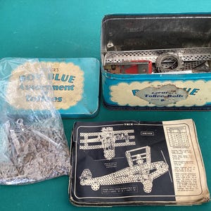Può includere: Una scatola di metallo blu e bianca con il testo "Royal Blue Assortment Toffee Rolls" sul coperchio. La scatola è aperta e contiene una varietà di pezzi di metallo. Una busta di plastica trasparente con pezzi di metallo è accanto alla scatola. È inclusa anche una scheda di istruzioni in bianco e nero con un diagramma di un biplano.