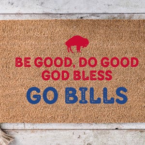 Può includere: Un zerbino rettangolare in fibra di cocco con la scritta "BE GOOD, DO GOOD, GOD BLESS, GO BILLS" in rosso e blu. Un logo di bufalo rosso è in alto. Il tappeto è su una superficie di legno chiaro.