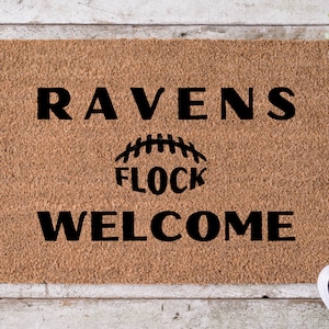May include: A brown coir doormat with the text "RAVENS FLOCK WELCOME" in black lettering. The text is centered on the mat and the word "FLOCK" is above a stylized football.