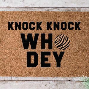 Op de afbeelding: Een bruine kokosvezel deurmat met de tekst "Knock Knock Who Dey" in zwarte letters. Een zwarte en bruine zebraprint cirkel bevindt zich naast het woord "Who".