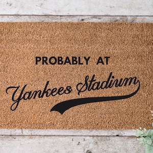 以下が含まれることがあります： 天然ココナッツ繊維で作られた長方形のドアマット。「PROBABLY AT Yankees Stadium」というテキストが黒色で表示され、「Stadium」の文字は筆記体です。背景は明るい茶色です。