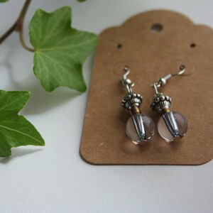 May include: A pair of silver earrings with clear glass beads and ornate silver accents. The earrings are displayed on a brown card.