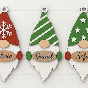 May include: Three wooden Christmas gnome ornaments. Each gnome has a different colored hat: red with a snowflake, green with white stripes, and green with white stars. Each ornament has a name: Olivia, Daniel, and Sofia.