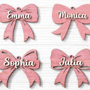 Può includere: Quattro ornamenti a forma di fiocco rosa in legno con i nomi Emma, Monica, Sophia e Julia. I nomi sono in bianco, con un carattere corsivo. Ogni fiocco ha un anello per appenderlo.