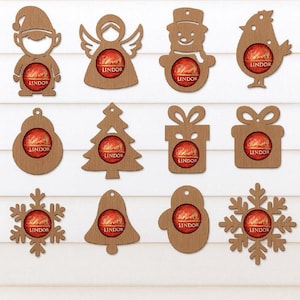 Peut inclure: Diverses décorations de Noël en bois, comprenant un bonhomme de neige, un ange et un flocon de neige. Chaque ornement contient un chocolat Lindor dans une découpe circulaire, avec le logo Lindor. Les décorations sont en bois brun clair.