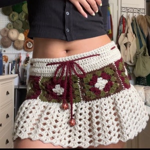 Puede incluir: Una falda de crochet blanca con una cintura estampada en color burdeos y verde. La falda tiene un dobladillo con volantes y est&aacute; atada con una cinta de color burdeos.