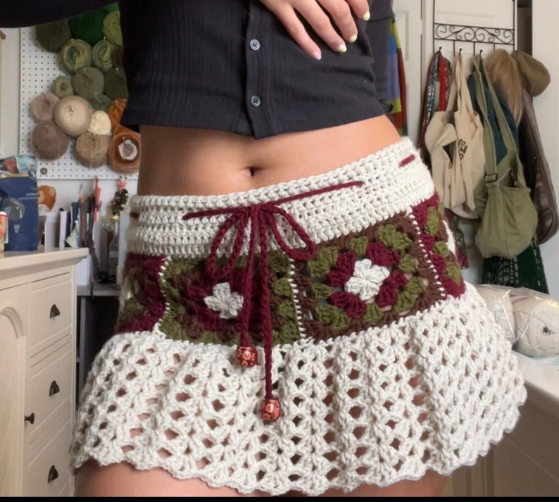 Puede incluir: Una falda de crochet blanca con una cintura estampada en color burdeos y verde. La falda tiene un dobladillo con volantes y est&aacute; atada con una cinta de color burdeos.