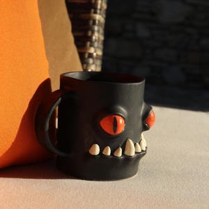 Pode incluir: Caneca de cerâmica preta com um design de rosto de monstro. A caneca apresenta olhos grandes laranja, pequenas protuberâncias no nariz laranja e dentes brancos. A caneca tem uma alça curva.