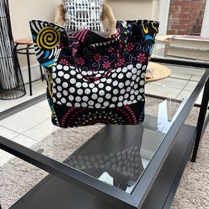 Può includere: Una borsa tote nera e bianca con un motivo a pois e disegni geometrici colorati. La borsa ha due manici neri ed è realizzata in tessuto.