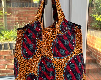 Pode incluir: Uma sacola estampada com um design vibrante. A sacola apresenta um padrão repetitivo semelhante a paisley em vermelho, azul marinho e verde-azulado em um fundo laranja com bolinhas pretas. A sacola tem uma alça combinando e está pendurada em uma porta branca.